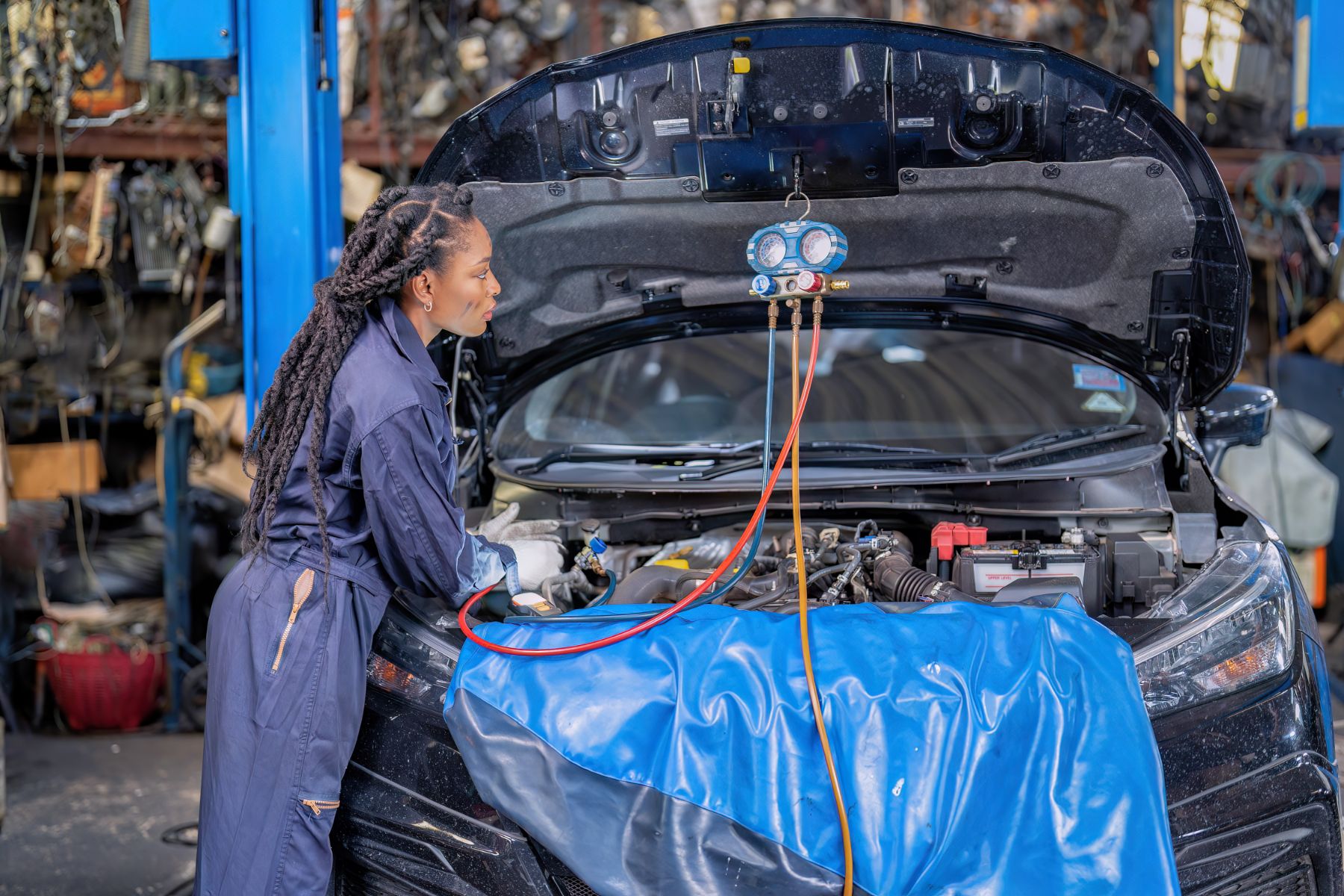 A mechanic in coveralls checks the engine of a car with its hood open, using diagnostic equipment and hoses while standing in an auto repair shop.