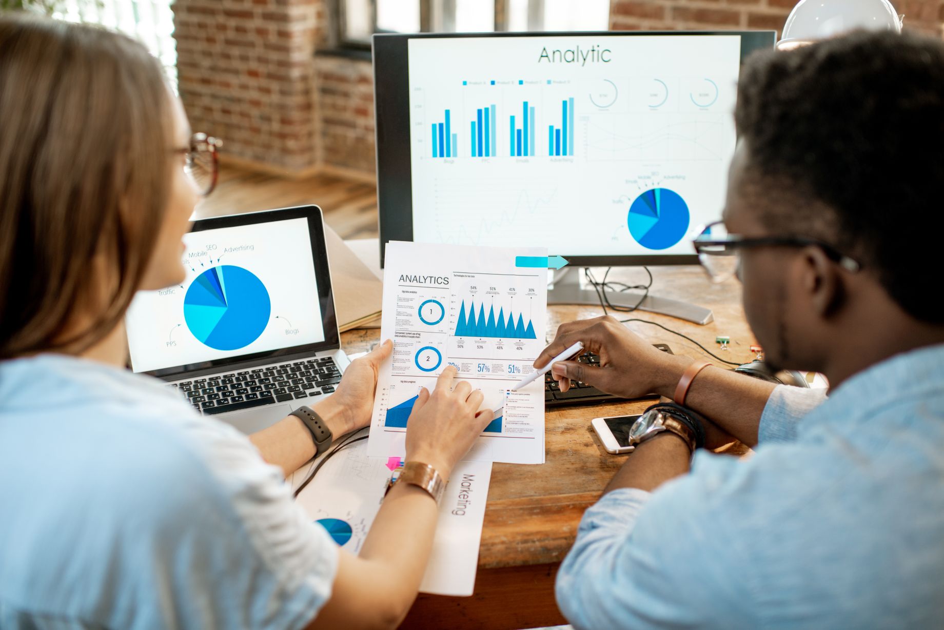 Two people review marketing analytics charts at a desk with a laptop and monitors displaying blue graphs and data visualizations.