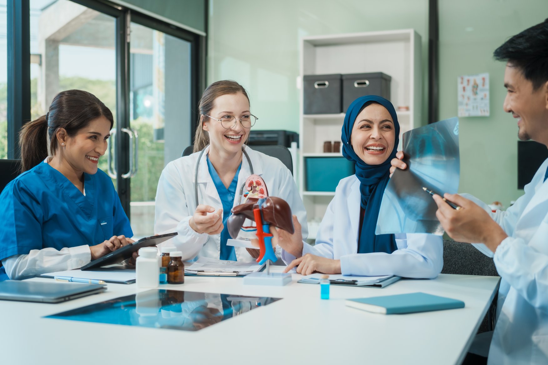 Four medical professionals in lab coats and scrubs sit at a table examining X-rays and an anatomical model, smiling and discussing together in a bright office.