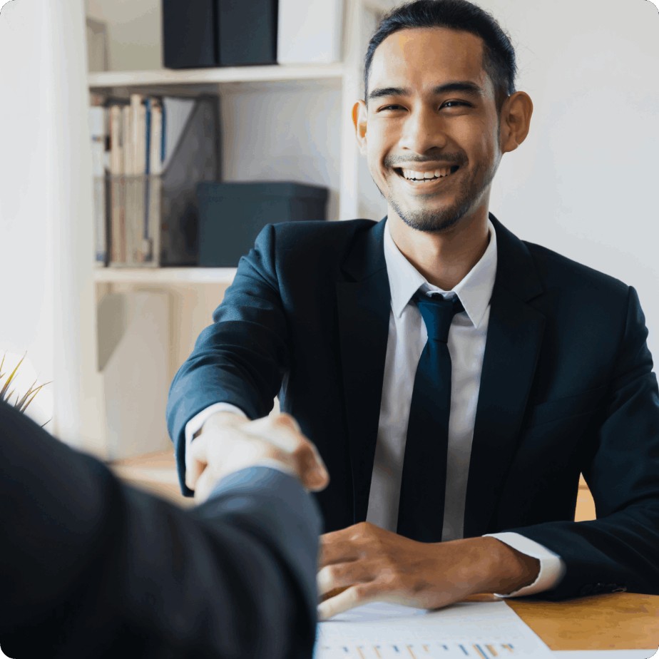 A man in a suit is smiling and shaking hands with another person across a desk in an office setting.
